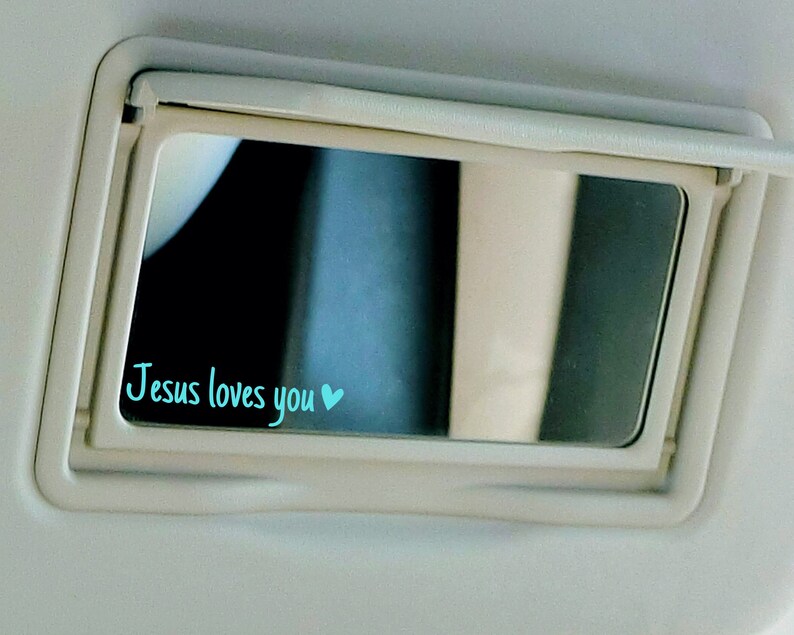 May include: A white car visor with a mirror that has the words "Jesus loves you" written in blue with a small blue heart.