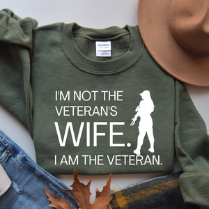 May include: Green sweatshirt with white text that reads "I'm not the veteran's wife. I am the veteran." A white silhouette of a woman holding a rifle is in the centre of the text.
