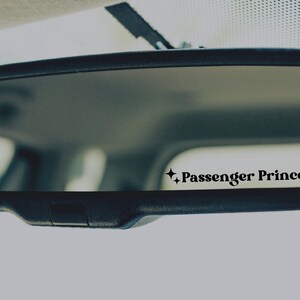 Op de afbeelding: Een achteruitkijkspiegel met een zwarte sticker waarop "Passenger Princess" staat met twee sterren aan weerszijden.