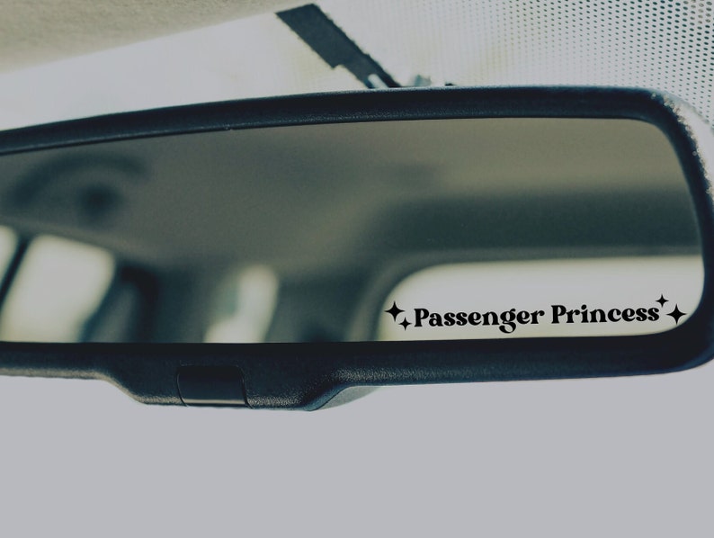 Op de afbeelding: Een achteruitkijkspiegel met een zwarte sticker waarop "Passenger Princess" staat met twee sterren aan weerszijden.