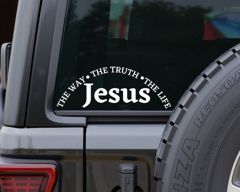 Puede incluir: Calcoman&iacute;a de vinilo blanco con el texto "Jes&uacute;s" en una fuente grande y el texto "THE WAY THE TRUTH THE LIFE" en una fuente m&aacute;s peque&ntilde;a, curvado alrededor de la palabra "Jes&uacute;s". La calcoman&iacute;a est&aacute; en la ventana trasera de un coche negro.