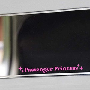 Op de afbeelding: Een zwarte rechthoekige spiegel met een zilveren frame. De spiegel heeft een roze sticker met de tekst "Passenger Princess" en twee kleine sterren aan weerszijden van de tekst.