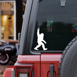 May include: White vinyl sticker of a Bigfoot silhouette giving the peace sign. The sticker is on the back window of a red SUV.