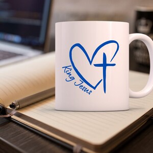 May include: White ceramic mug with a blue heart and cross design. The text "King Jesus" is written inside the heart.