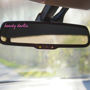 May include: A black rearview mirror with an auto-dimming feature. The mirror has a small yellow light and the word "AUTO" printed on it. The mirror is mounted in a car and the reflection shows a blurry image of trees and a road.