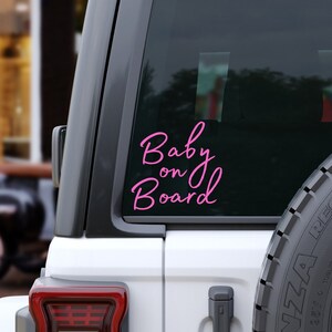 K&ouml;nnte beinhalten: Ein pinkfarbener Vinyl-Aufkleber mit der Aufschrift "Baby on Board" in Schreibschrift ist auf der Heckscheibe eines wei&szlig;en Fahrzeugs angebracht. Der Aufkleber ist vor dem dunkel get&ouml;nten Glas zu sehen, ein Teil des Reserverads ist sichtbar.
