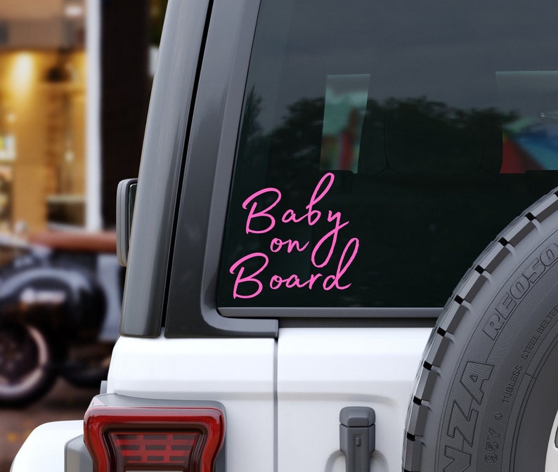 K&ouml;nnte beinhalten: Ein pinkfarbener Vinyl-Aufkleber mit der Aufschrift "Baby on Board" in Schreibschrift ist auf der Heckscheibe eines wei&szlig;en Fahrzeugs angebracht. Der Aufkleber ist vor dem dunkel get&ouml;nten Glas zu sehen, ein Teil des Reserverads ist sichtbar.