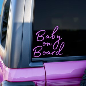 K&ouml;nnte beinhalten: Ein rosa Fahrzeug mit einem schwarzen Fenster, das einen hellvioletten "Baby on Board"-Aufkleber in Schreibschrift zeigt. Das Bild zeigt einen Teil des Fahrzeug&auml;u&szlig;eren, einschlie&szlig;lich eines Reifens und einen Blick auf eine Palme.