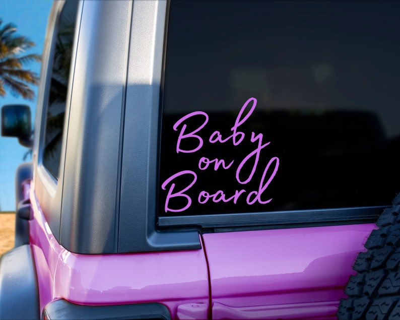 K&ouml;nnte beinhalten: Ein rosa Fahrzeug mit einem schwarzen Fenster, das einen hellvioletten "Baby on Board"-Aufkleber in Schreibschrift zeigt. Das Bild zeigt einen Teil des Fahrzeug&auml;u&szlig;eren, einschlie&szlig;lich eines Reifens und einen Blick auf eine Palme.