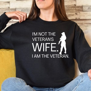 May include: Black sweatshirt with white text that reads "I'm not the veteran's wife. I am the veteran."  A white silhouette of a woman holding a rifle is to the right of the text.