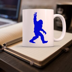 May include: White ceramic coffee mug with a blue silhouette of a Bigfoot giving the peace sign.
