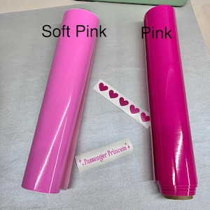 Op de afbeelding: Twee rollen vinyl-plakfolie, &eacute;&eacute;n in zacht roze en &eacute;&eacute;n in roze. De rollen zijn gelabeld met "Soft Pink" en "Pink". De afbeelding bevat ook een vel met roze hart stickers en een sticker met de tekst "Passenger Princess".