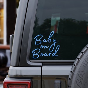 K&ouml;nnte beinhalten: Ein hellblauer "Baby on Board" Aufkleber ist auf der Heckscheibe eines grauen Fahrzeugs angebracht. Der kursive Text hebt sich vom dunklen Glas ab. Das Bild zeigt einen Teil des Fahrzeug&auml;u&szlig;eren, einschlie&szlig;lich eines Reifens.