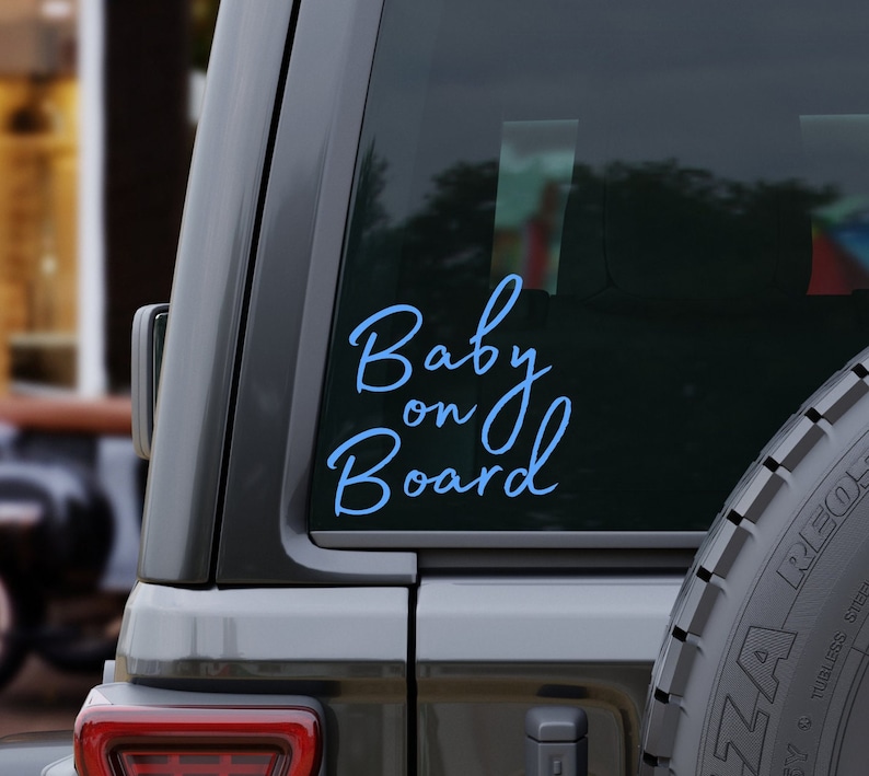 K&ouml;nnte beinhalten: Ein hellblauer "Baby on Board" Aufkleber ist auf der Heckscheibe eines grauen Fahrzeugs angebracht. Der kursive Text hebt sich vom dunklen Glas ab. Das Bild zeigt einen Teil des Fahrzeug&auml;u&szlig;eren, einschlie&szlig;lich eines Reifens.
