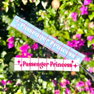 Op de afbeelding: Twee witte vinyl stickers met de tekst "Passenger Princess" in roze letters. De stickers zitten op een geruit achterblad en worden met een pincet van de ondergrond gehaald.