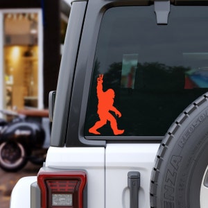 May include: Orange vinyl sticker of a Bigfoot silhouette making a peace sign on a white car. The sticker is on the back window of the car.