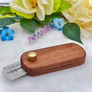 May include: A wooden razor blade holder with a brass knob. The holder is open and a silver razor blade is visible with the text "FACON PLATINUM STAINLESS" printed on it.