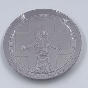 May include: A circular gray ceramic plate featuring a raised relief design of a central figure with arms outstretched, wrapped in cloth, set against a cross. A star appears in the upper right, and the text 19-KING OF KINGS-70 is embossed.