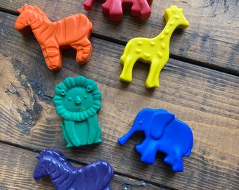 May include: A set of six animal-shaped wax crayons in various colors, including a red elephant, an orange zebra, a yellow giraffe, a green lion, a blue elephant, and a purple zebra, all arranged on a dark brown wooden surface.