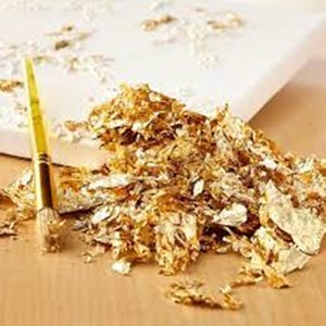 Può includere: Un mucchio di scaglie di foglia d'oro su una superficie di legno. Un pennello è sdraiato accanto alle scaglie.