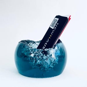 May include: A teal blue resin business card holder shaped like a wave with a black business card partially submerged in the wave.