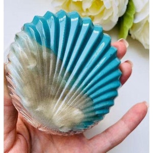 May include: A seashell-shaped soap bar with a turquoise blue and beige color scheme. The soap has a ribbed texture and is held in a hand, with white flowers in the background. The soap is a decorative item.