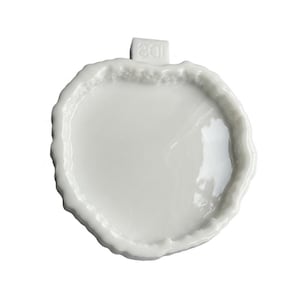 May include: A white ceramic dish shaped like an apple with a glossy finish and a textured, uneven rim. A small rectangular tab extends from the top, featuring the embossed text ID8. The dish has a smooth, concave center and a solid white color.