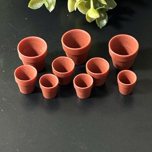 Może przedstawiać: Kolekcja małych, terakotowych glinianych doniczek o różnych rozmiarach. Doniczki są ułożone w grupie, z kilkoma większymi i kilkoma mniejszymi. Zielone liście widoczne w tle.