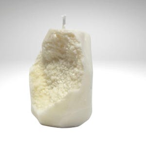Op de afbeelding: Een witte kaars in de vorm van een rots met een ruw, getextureerd oppervlak. De kaars heeft een witte lont en is gemaakt van natuurlijke was.