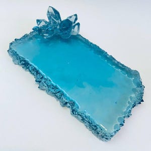 Puede incluir: Una bandeja rectangular turquesa con un grupo de cristales. La bandeja tiene un borde texturizado y dentado y una superficie lisa y brillante. El grupo de cristales está hecho de cristales claros y facetados.