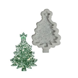 Puede incluir: Un adorno de árbol de Navidad verde brillante con una estrella, junto a un molde de silicona blanco con forma de árbol de Navidad. El adorno está hecho de muchas piezas pequeñas y reflectantes.