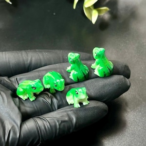 Könnte beinhalten: Eine Sammlung von sechs kleinen, grünen Froschfiguren mit unterschiedlichen Farbtönen und Mustern. Die Frösche befinden sich in verschiedenen Posen, einige sitzen und andere stehen. Die Figuren werden auf einer schwarzen Oberfläche präsentiert.