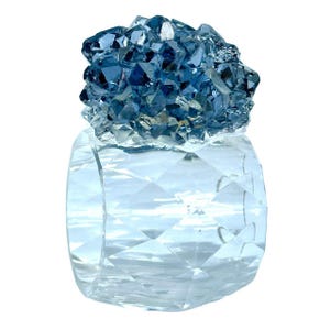 May include: A clear, faceted glass napkin ring with a cluster of blue crystals on top. The crystals have a rough, irregular texture, and the glass ring has a geometric pattern. The overall effect is elegant and decorative.