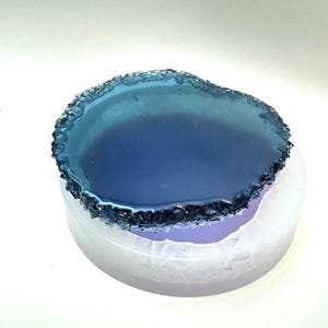 Può includere: Un sottobicchiere in resina di agata blu chiaro con uno stampo in silicone bianco. Il sottobicchiere ha un bordo ruvido e naturale.
