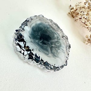 May include: A circular resin coaster featuring a swirling dark gray and white smoke-like pattern in the center, surrounded by a thick, clear, faceted border that mimics the appearance of jagged, crystalline rock edges with a glossy finish.