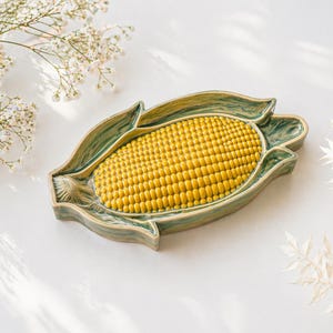 以下が含まれることがあります： 黄色いトウモロコシのデザインが施された、トウモロコシ型のセラミック製サービングディッシュ。縁には緑と青の大理石模様があり、トウモロコシの殻に似ています。白い表面に置かれています。