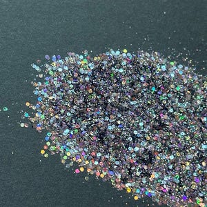 Pode incluir: Uma pilha de glitter iridescente com uma mistura de cores, incluindo prata, roxo e azul. As partículas de glitter são de vários tamanhos e formas, refletindo a luz. O fundo é uma superfície cinza escuro.