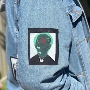 May include: Light blue denim jacket with a gray hood. The jacket features a black and white graphic patch on the sleeve and a larger patch on the back with a green and red design.
