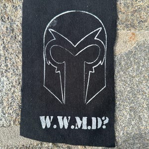 May include: Black fabric patch featuring a white outline of a helmet-like design. Below the graphic, the text "W.W.M.D.?" is printed in white. The patch is rectangular and appears to be made of a woven material.