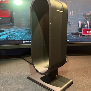 May include: A black headphone stand with a rectangular base and a curved, U-shaped top. The stand is made of a matte material and is designed to hold headphones. The stand is on a dark surface.