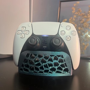 May include: A white and black PlayStation 5 DualSense controller with a teal honeycomb-patterned stand. The controller has a touchpad, directional buttons, and action buttons. The stand is designed to hold the controller upright.