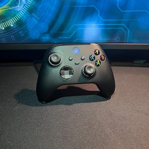 May include: A black Xbox controller with a central directional pad, two analog sticks, and colored action buttons. The controller has a matte finish and is positioned in front of a computer monitor with a blue and white graphic.