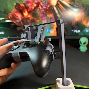 May include: A black gaming controller with gray attachments is held in front of a computer screen. A white and gray stand is attached to the controller. A small green alien figurine is in the background.