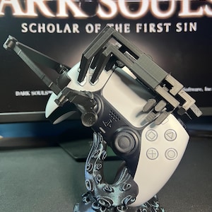 May include: A black and white PlayStation 5 controller with a custom black grip attachment, resting on a decorative stand. The stand has an octopus-like design. The background displays the text "DARK SOULS SCHOLAR OF THE FIRST SIN."