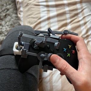 Puede incluir: Mando Xbox negro con un accesorio gris impreso en 3D. El mando tiene botones de colores y una cruceta. El accesorio parece estar diseñado para mejorar la funcionalidad del mando.
