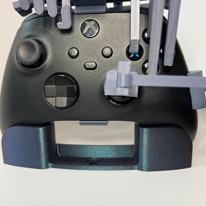 May include: Black Xbox controller with a custom gray 3D-printed attachment. The controller has a matte finish with the Xbox logo and various buttons. The attachment appears to be designed to hold the controller.