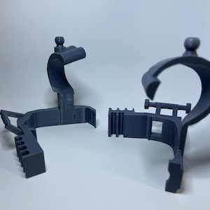 May include: Two dark gray 3D-printed plastic components. One component has a curved, circular shape with a small ball on top. The other component has a rectangular shape with a series of vertical slots. Both components are designed to fit together.