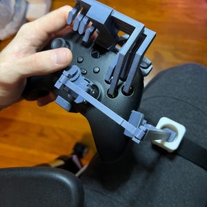 May include: A black Xbox controller with a custom 3D-printed gray attachment. The attachment appears to be designed to assist with gaming. The controller is connected to a strap worn on the leg.