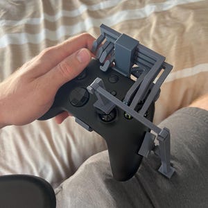 May include: A black Xbox controller with a gray 3D-printed attachment. The attachment appears to be designed to hold the controller. The controller is being held by a person.