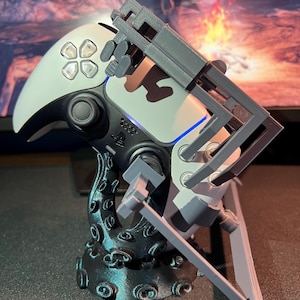 May include: A white and black video game controller with a gray, articulated attachment. The controller is mounted on a black, tentacle-shaped base. The attachment has a mechanical design.